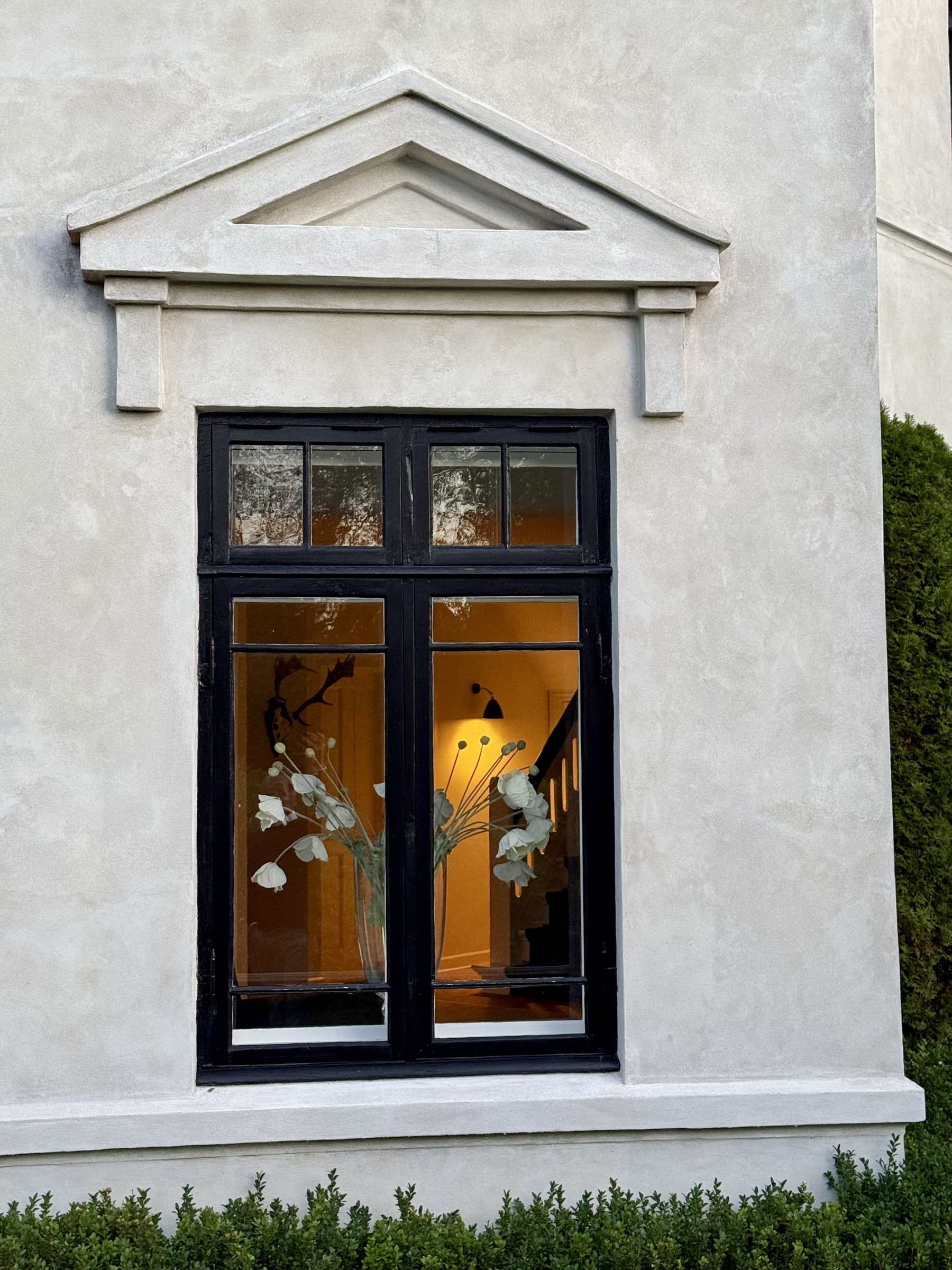 Black-framed window beneath a classical pediment, warm light and white flowers inside
